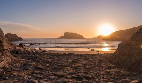Golden light from the rising sun reflects across the wet sand and coastal rocks of Arnia Beach,