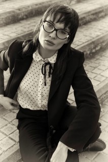 A stylish woman wearing glasses and a vintage-inspired suit sits on stone steps. Captured in black