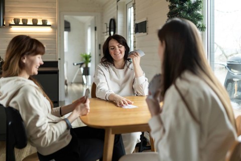Three friends gather around a wooden table, smiling and engaged in a card game. The cozy interior