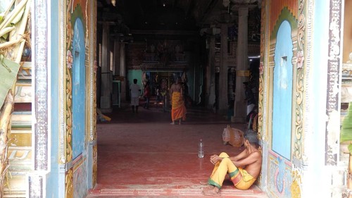 A man sits inside a colorful temple while others move in the background, Sri Lanka