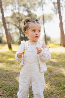 A young girl stands in a sunny forest, holding a small yellow flower. She wears a white knit