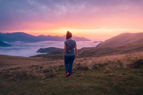 A woman stands on a grassy hill, gazing at a stunning sunset over rolling mountains and clouds in