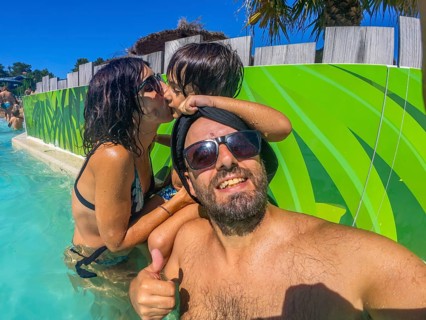 Family taking a joyful selfie in a sunny water park pool, parents and child smiling and kissing,