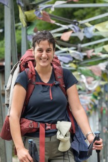 Smiling woman with a backpack and trekking gear stands on a path adorned with prayer flags, ready