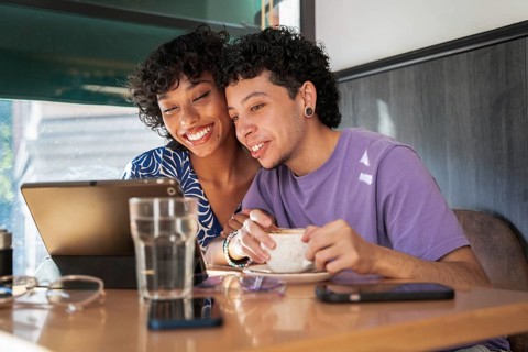 A joyful multiethnic LGBTQ+ couple sits closely in a cozy café, sharing a warm moment over coffee