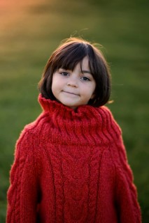 A young girl with a joyful expression, wearing a red cable-knit sweater, stands outdoors with a