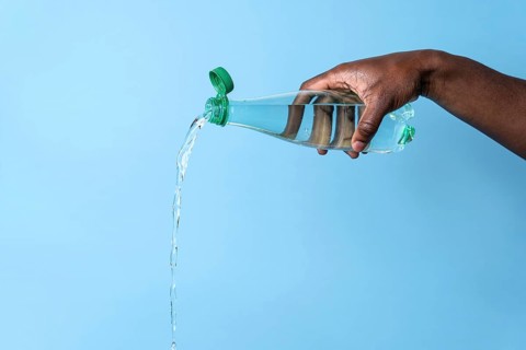 A close-up of a hand pouring water from a clear plastic bottle against a light blue background. The