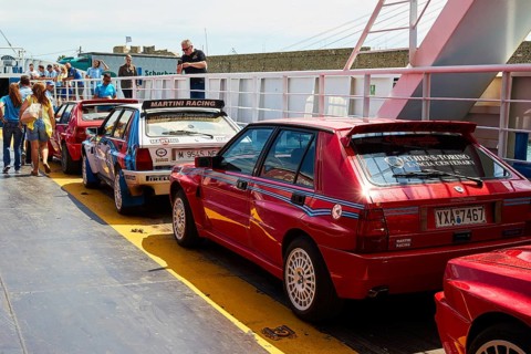 Several red vintage cars on a ferry surrounded by people under a bright sky, rally cars,