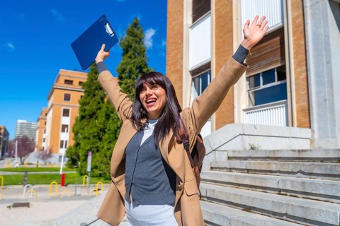Happy young woman raising arms in front of a university building, celebrating a success or
