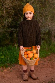 Young child dressed in autumnal clothing holds a basket filled with fresh oranges. The scene is set