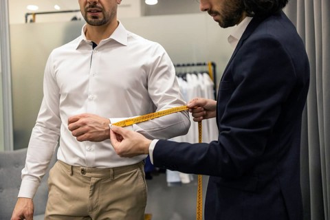 A tailor measures a client sleeve length with a yellow tape in a suit store. The client wears a