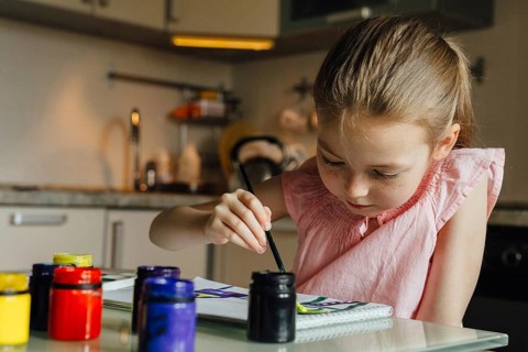 A young girl concentrates on painting with vibrant watercolors, surrounded by various paint jars on