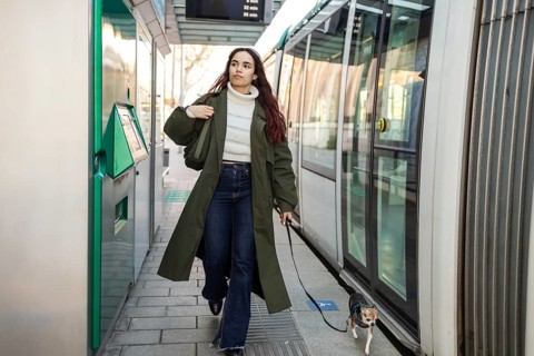 Woman walks her chihuahua at a tram stop, wearing a green coat and jeans. Urban setting with public