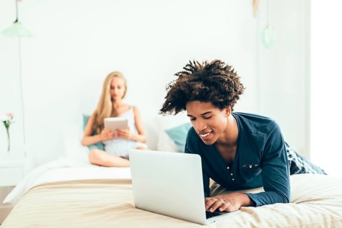 A young man lies on a bed using a laptop, while a woman sits in the background with a tablet. The