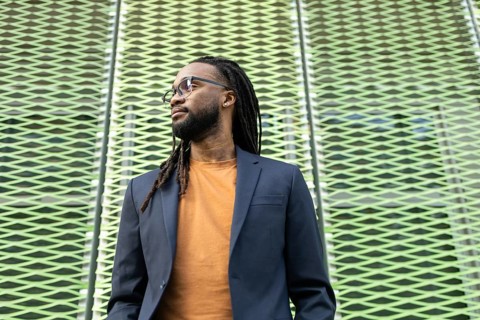 A cuban business man with long hair in glasses and a suit stands against a textured, patterned