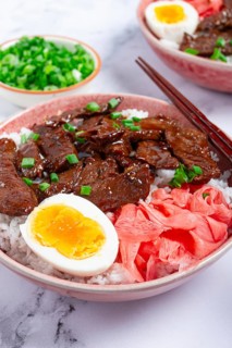 Gyudon Japanese beef with rice, pickled ginger, boiled egg slice, garnished with green onions, in a