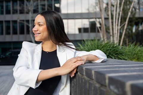 A smiling Hispanic woman in a white blazer enjoys a break outside a modern office building,