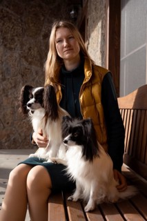 A woman wearing a yellow vest is seated on a wooden bench, accompanied by two purebred Papillon