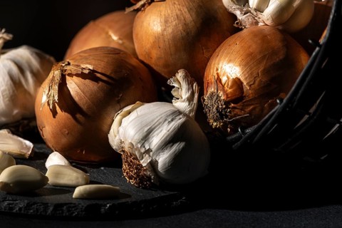 Onions and garlic next to a basket, dramatically lit on a dark background