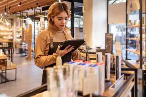 A cosmetics store worker in a brown sweater and black apron uses a tablet to check inventory. The