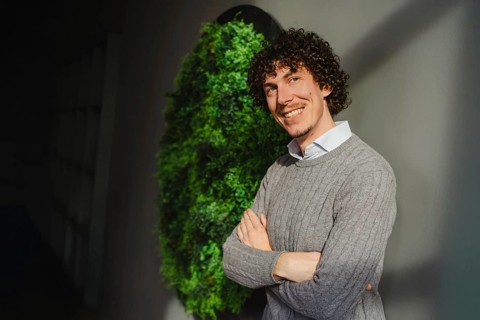A young man with curly hair stands in front of a green wall, symbolizing themes of ecology and