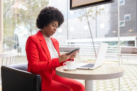 A woman in a red suit works on her phone and laptop in a modern cafe, reflecting a professional