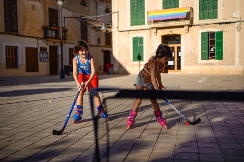 Two children play roller hockey with sticks and an orange ball in a sunlit urban square, creating a