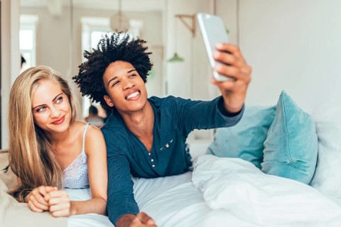 A cheerful couple is lying on a bed taking a selfie with a smartphone. The cozy bedroom has plush
