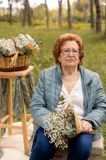 An elderly woman sits in an outdoor setting holding wildflowers with a serene expression. A wicker