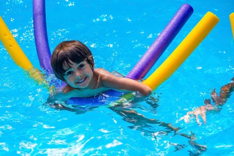 Young boy smiling and floating on colorful foam noodles in a clear blue pool, enjoying sunny summer