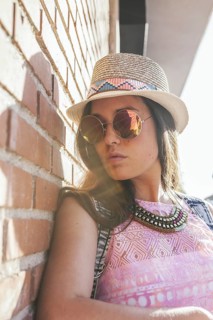 A fashionable woman leans against a brick wall, wearing a summer hat and sunglasses. Her pink