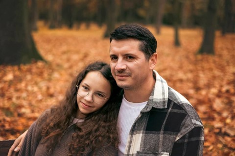 A father and daughter stand together in an autumn park, surrounded by vibrant fallen leaves. The