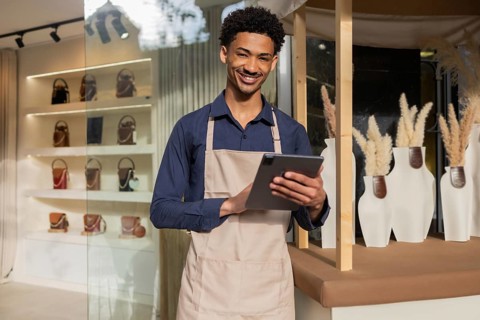 A cheerful store employee wearing an apron uses a tablet in a modern bag shop. Shelves display