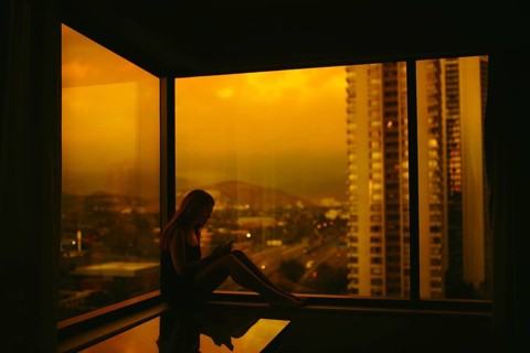 A woman is silhouetted against an urban skyline at sunset, browsing on her smartphone in a window