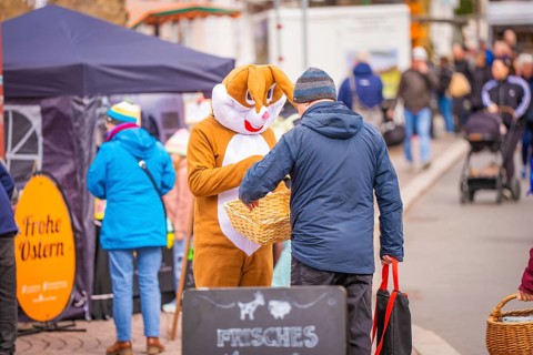 Bunny costume figure presents something to a man at a busy street market, Easter campaign,