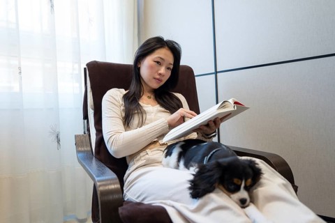 An Asian woman relaxes at home, sitting in a cozy chair by the window, engrossed in a book with her