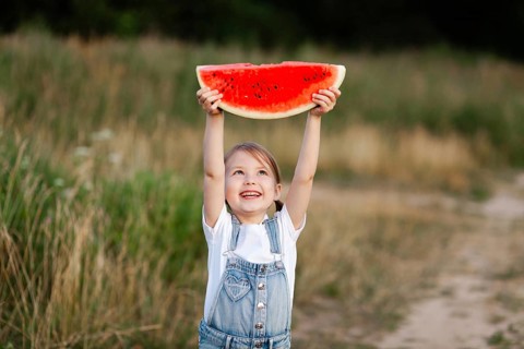A cheerful child holds up a large watermelon slice against a lush summer field backdrop, capturing