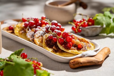 Delicious pancakes adorned with red currants are served on a white ceramic plate. They rest on a