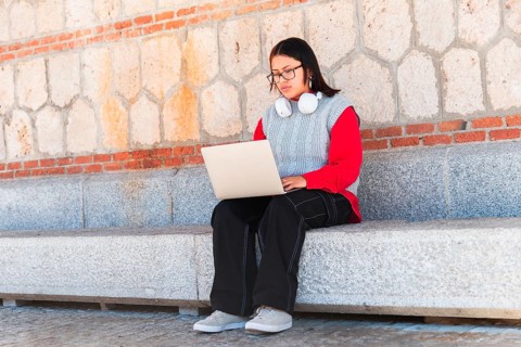 Hispanic student with glasses and headphones, focused on their laptop while seated on a stone bench