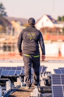 Man in work clothes standing between solar panels on a roof, with houses in the background, PV