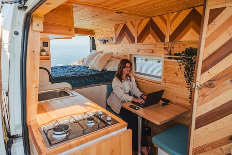 A woman works remotely on her laptop inside a cozy camper van, parked with a scenic view outside.