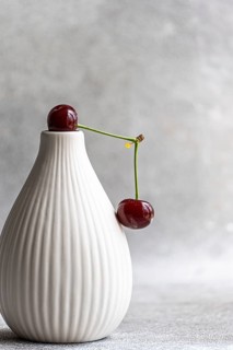 A ripe cherry delicately rests on an elegant white ceramic vase, with a soft concrete background