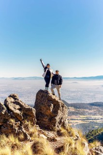 Two friends stand atop a rocky peak, enjoying a breathtaking mountain view under a clear blue sky