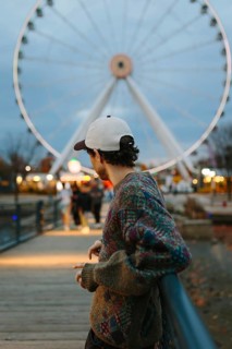 Side view of unrecognizable man wearing a cap and sweater, standing near a ferris wheel in
