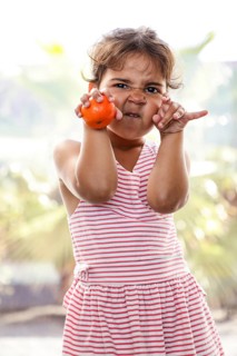 A child in a striped dress makes playful faces, holding a tangerine, enjoying a sunny day outdoors.