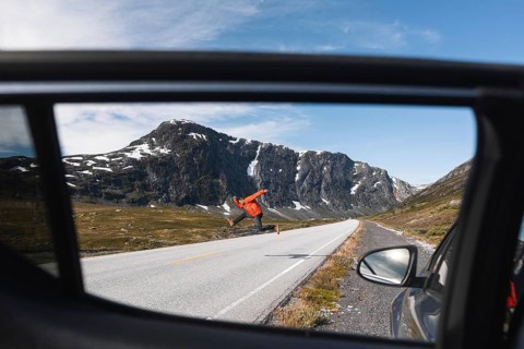 A traveler in a red jacket jumps joyfully on a mountain road in Norway, framed through a car window