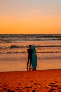 A man stands on the beach holding a blue surfboard. The sky is orange and the water is calm