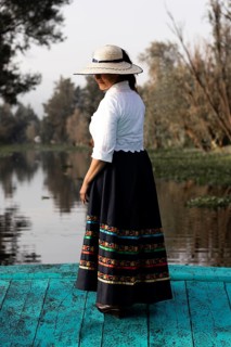 A woman in traditional clothing stands on a vibrant blue boat in Xochimilco, Mexico. The scenic