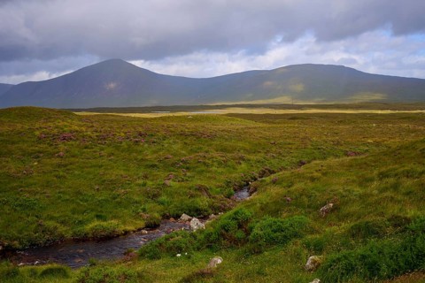 Green meadows and a flat valley with a small river surrounded by mountains under cloudy skies,