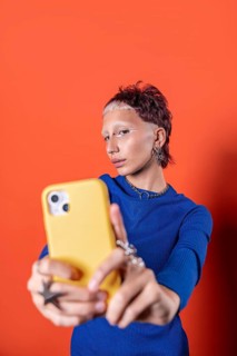 A stylish woman takes a selfie against a vivid orange backdrop, wearing a blue sweater and holding
